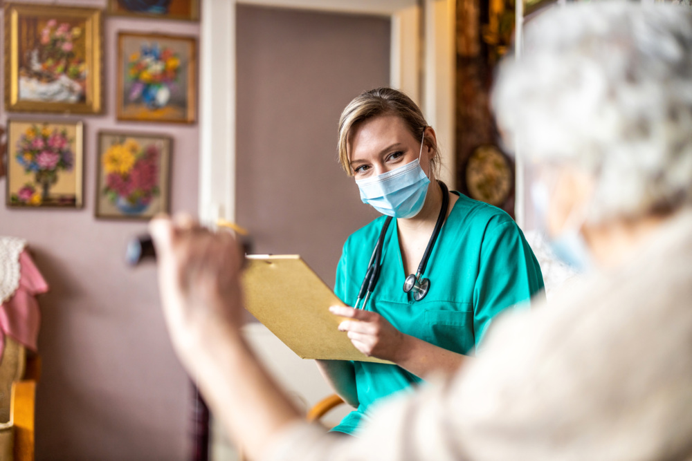Nurse in scrubs attending an older woman. Nurse is holding a clipboard and wearing a mask.